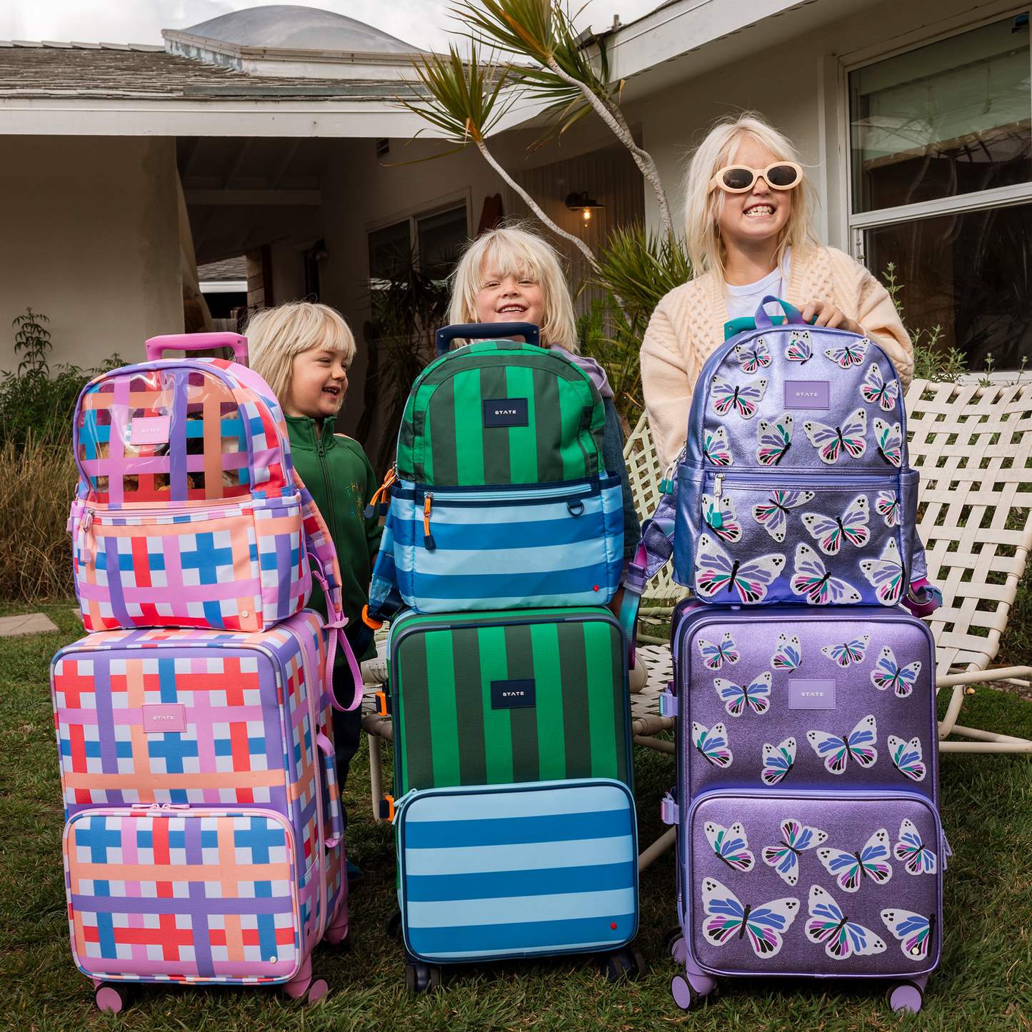 Two children and a woman standing behind colorful suitcases and backpacks on a grassy area.