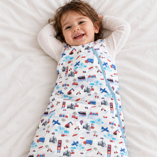 A smiling toddler lying on a white bed, wearing a zippered sleeping bag or swaddle with a colorful city-themed print of cars, buses, and airplanes.