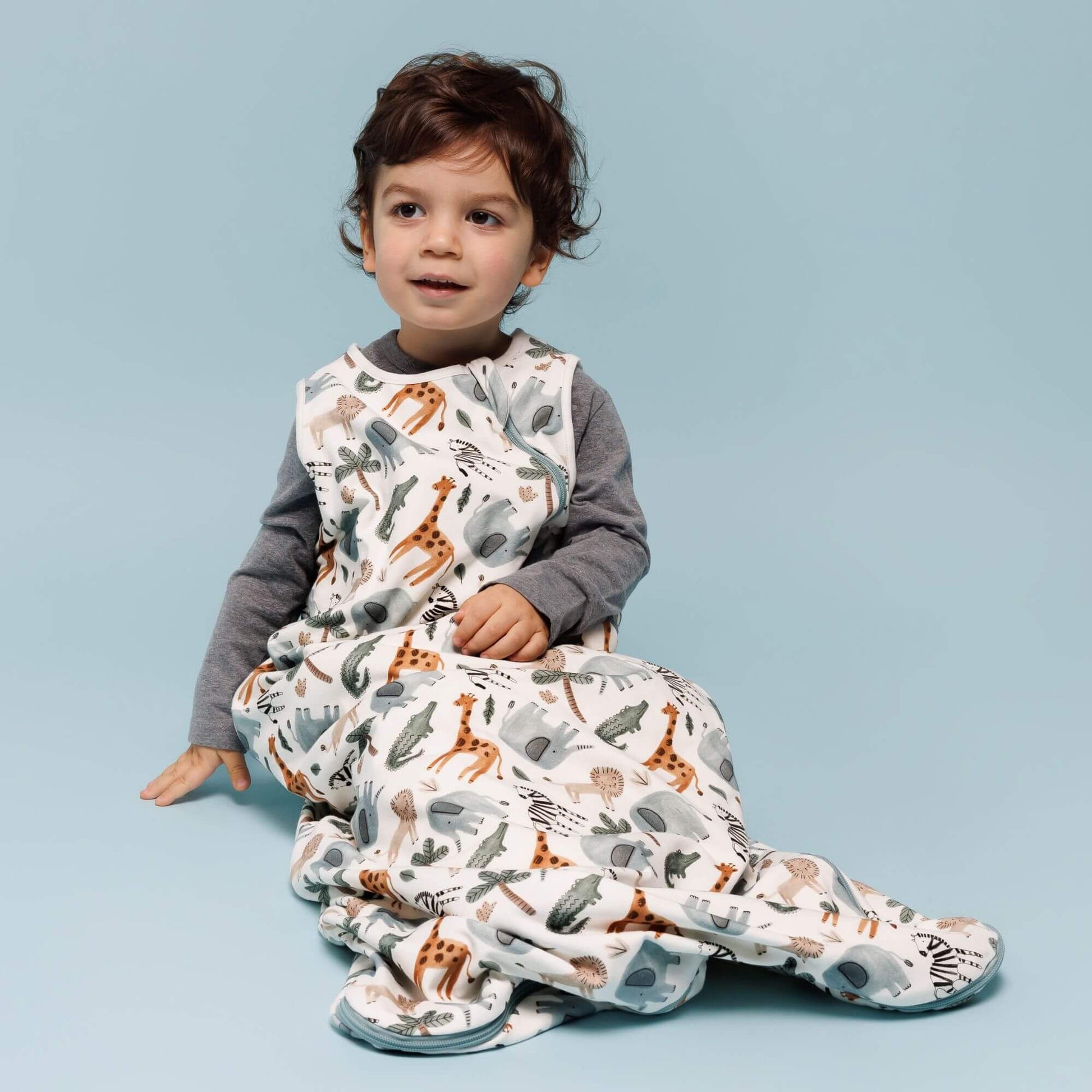 A young child smiling while sitting on a blue background, wearing the Norani Baby One & Done Sleep Sack in the "Safari Animals" print. The design showcases giraffes, elephants, and zebras, offering a playful and cozy sleep solution for toddlers.