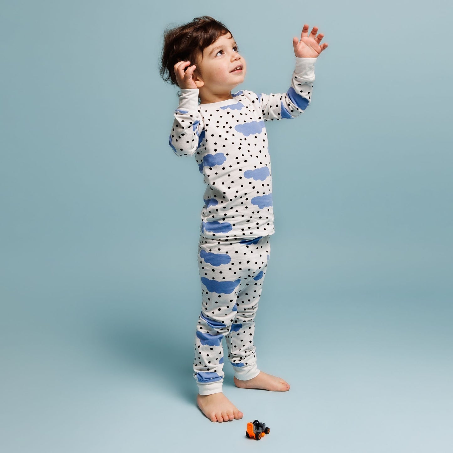 A toddler stands barefoot on a light blue background wearing Norani Baby's Blue Cloud Organic Pajamas, featuring a fun blue cloud and black polka dot pattern. The child looks upward with a curious and joyful expression, raising their arms slightly as if reaching or interacting with something unseen. A small orange toy car sits on the floor near their feet, emphasizing the comfort and playful design of Norani Baby's high-quality pajamas.