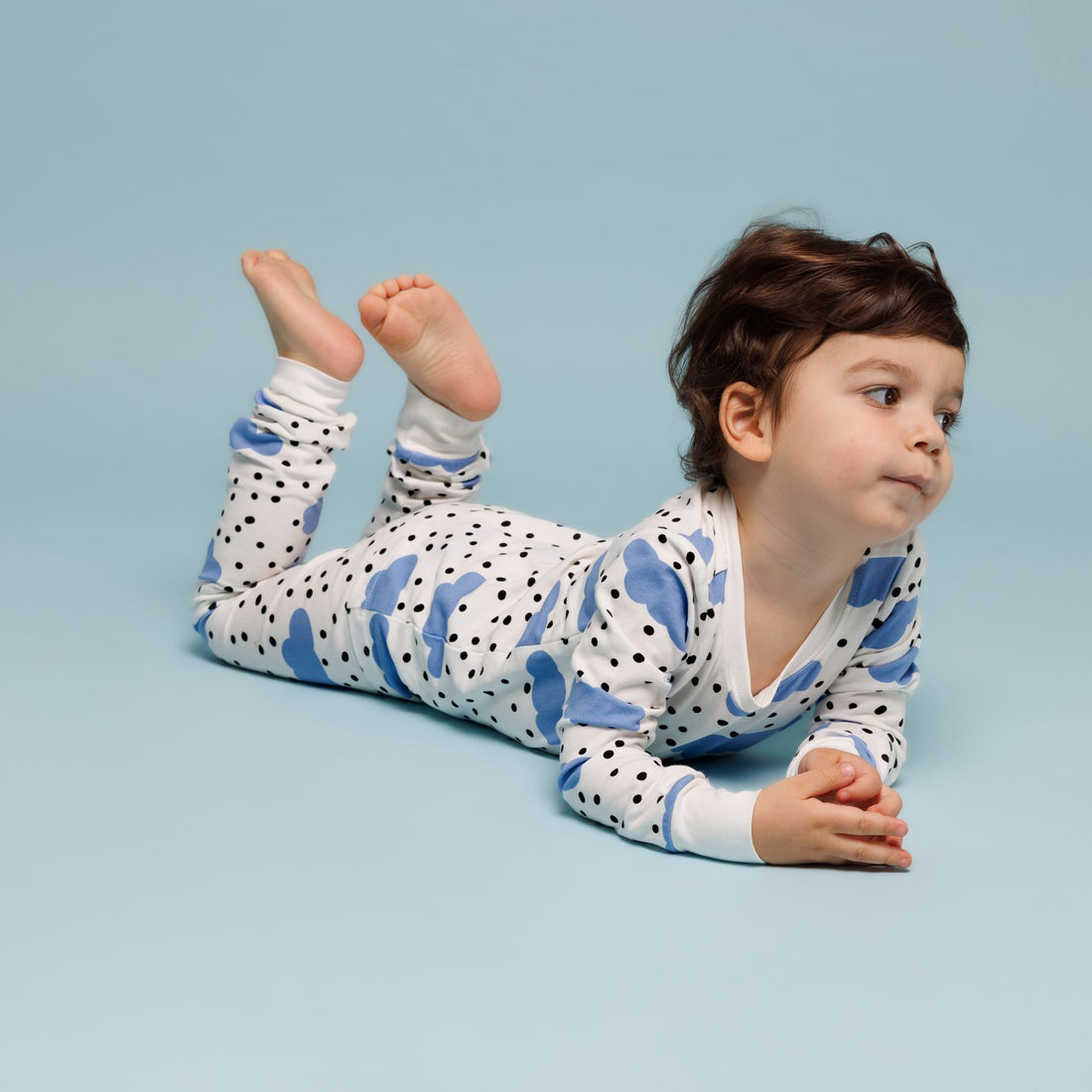 A toddler lounges on their stomach against a soft blue background, dressed in Norani Baby's Blue Cloud Organic Pajamas, showcasing cheerful blue cloud and black polka dot patterns. Their feet are playfully raised in the air, and they rest their hands together, gazing thoughtfully to the side. This relaxed pose highlights the softness and coziness of Norani Baby's organic sleepwear.