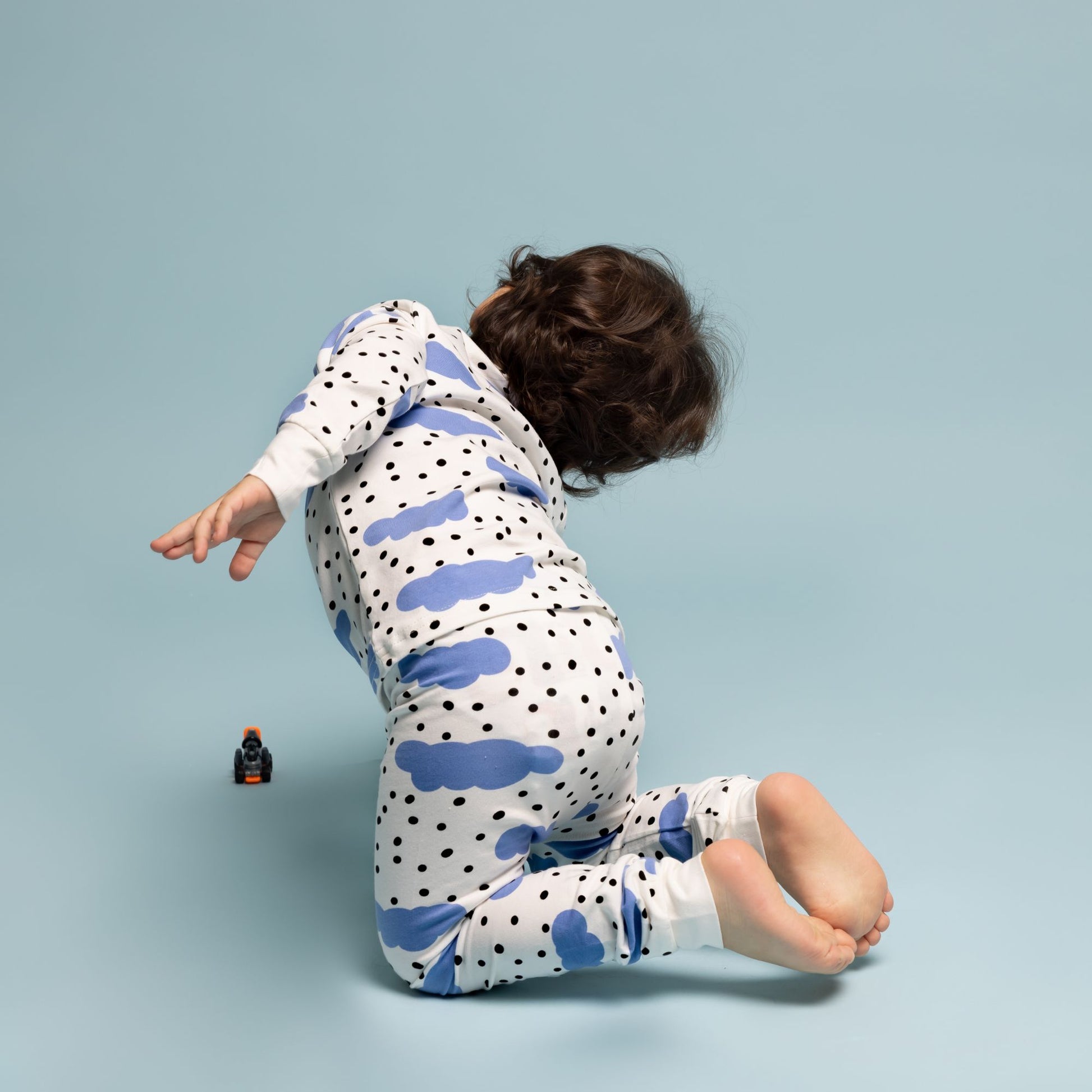 A toddler kneels on a light blue background wearing Norani Baby's Blue Cloud Organic Pajamas, featuring playful blue cloud and black polka dot patterns. They twist slightly, extending an arm in a moment of playful motion, while a small orange toy car sits nearby. The scene showcases the comfort and flexibility of Norani Baby's premium pajamas, perfect for active little ones.