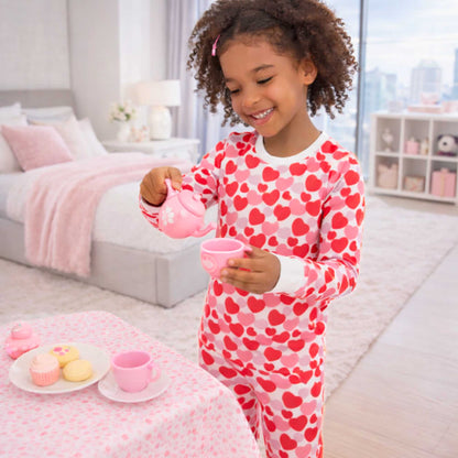 Child in heart-patterned pajamas by norani baby playing with a tea set in a bedroom.
