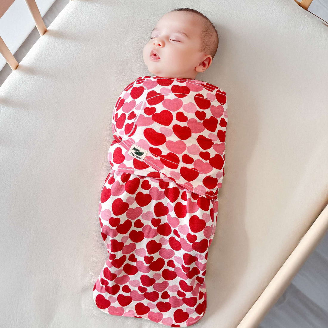 Baby swaddled in a red and pink heart-patterned blanket on a white surface.