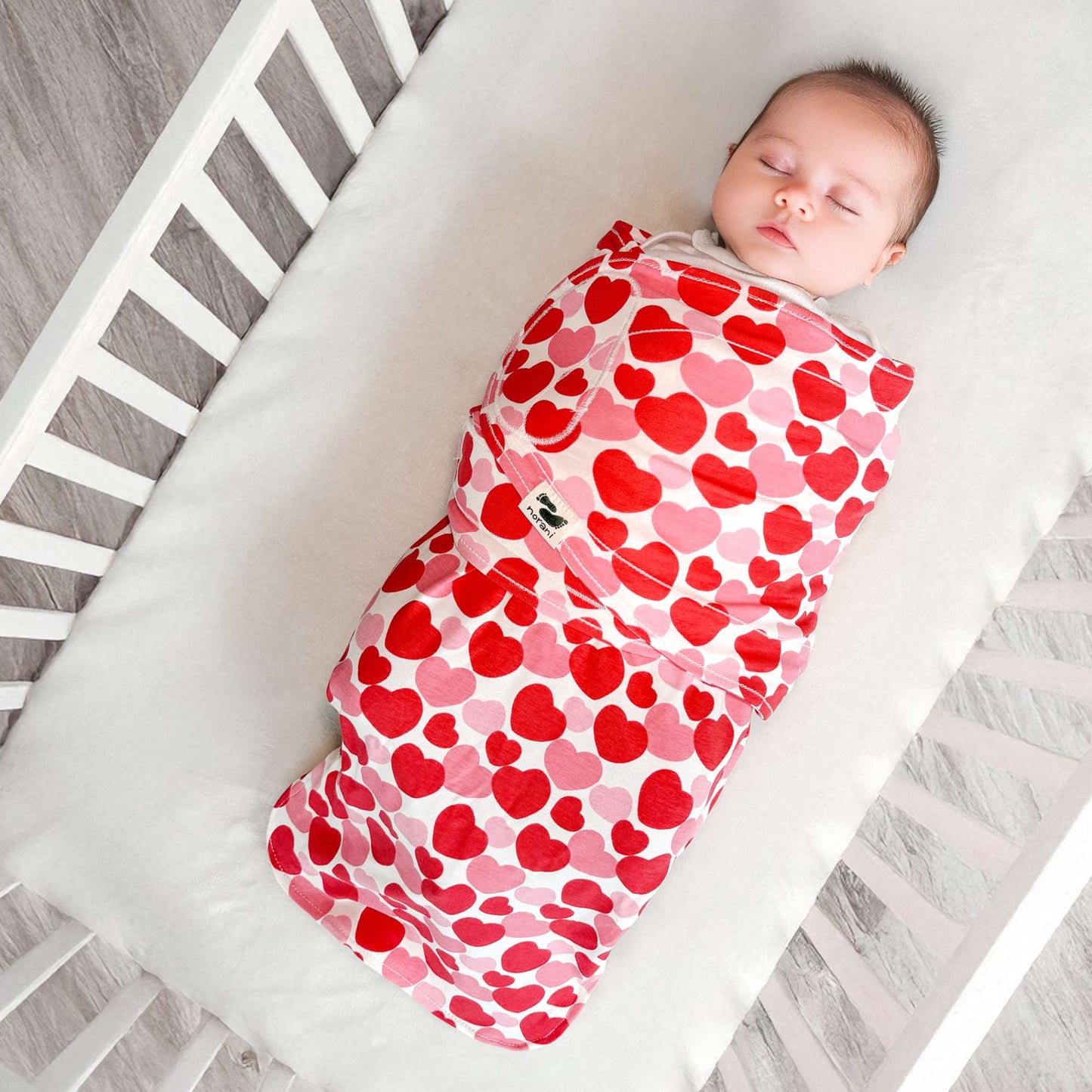 Baby swaddled in a red and pink heart-patterned blanket in a crib.