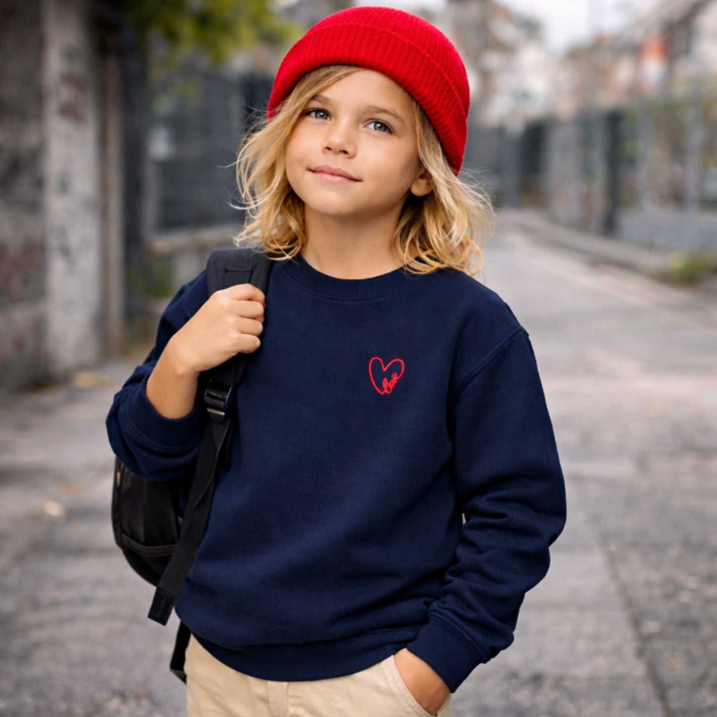 Child wearing a navy blue sweatshirt with a red heart design, red beanie, and black backpack on a street.