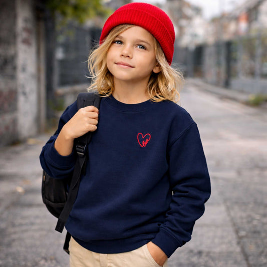 Child wearing a navy blue sweatshirt with a red heart design, red beanie, and black backpack on a street.