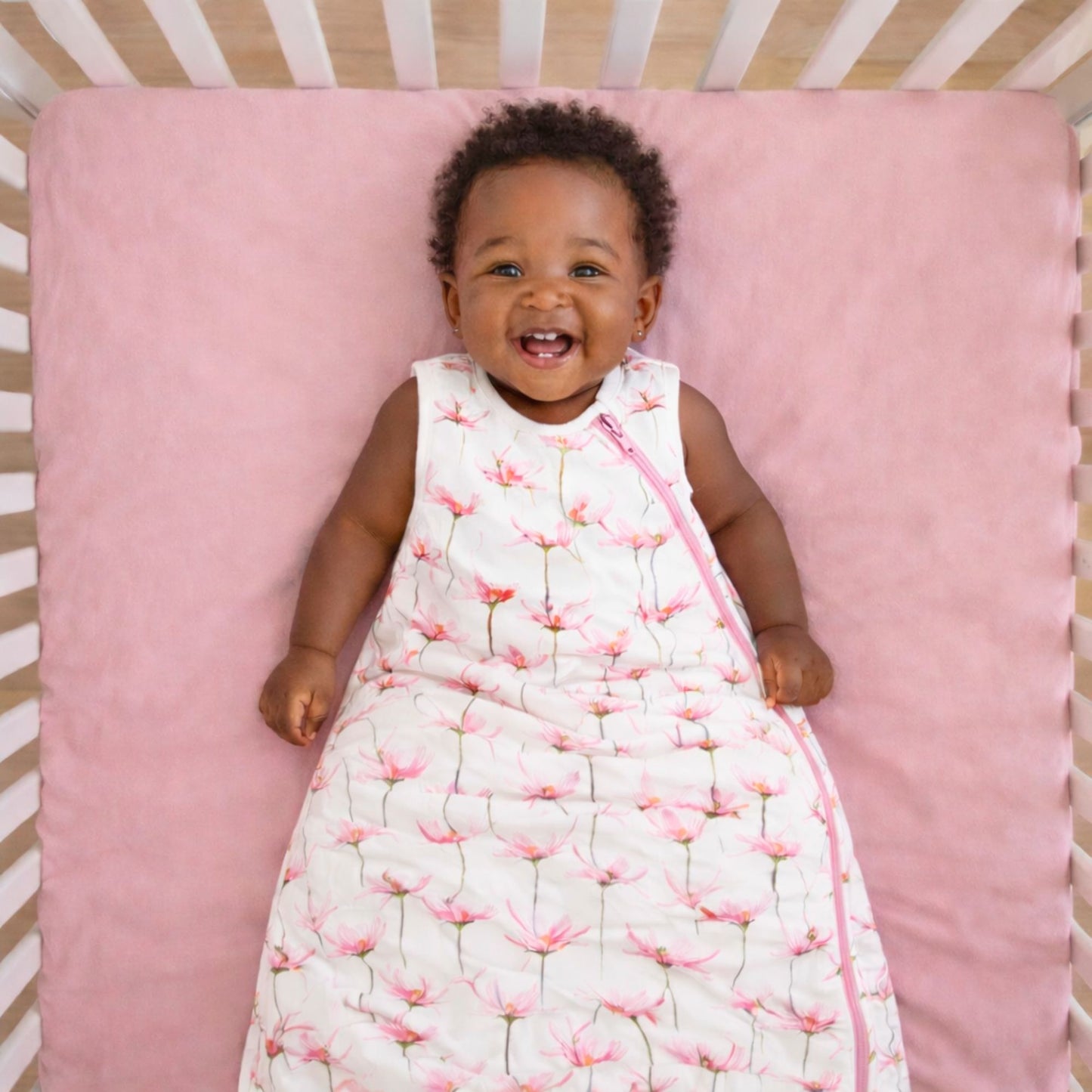 Baby lying on a pink mattress in a crib wearing the norani baby one & done sleep sack in pink petals
