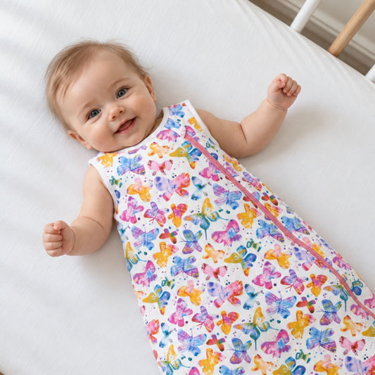 A baby lying on a white bed wearing a sleeveless sleeping bag or swaddle with a colorful butterfly print and a pink zipper.
