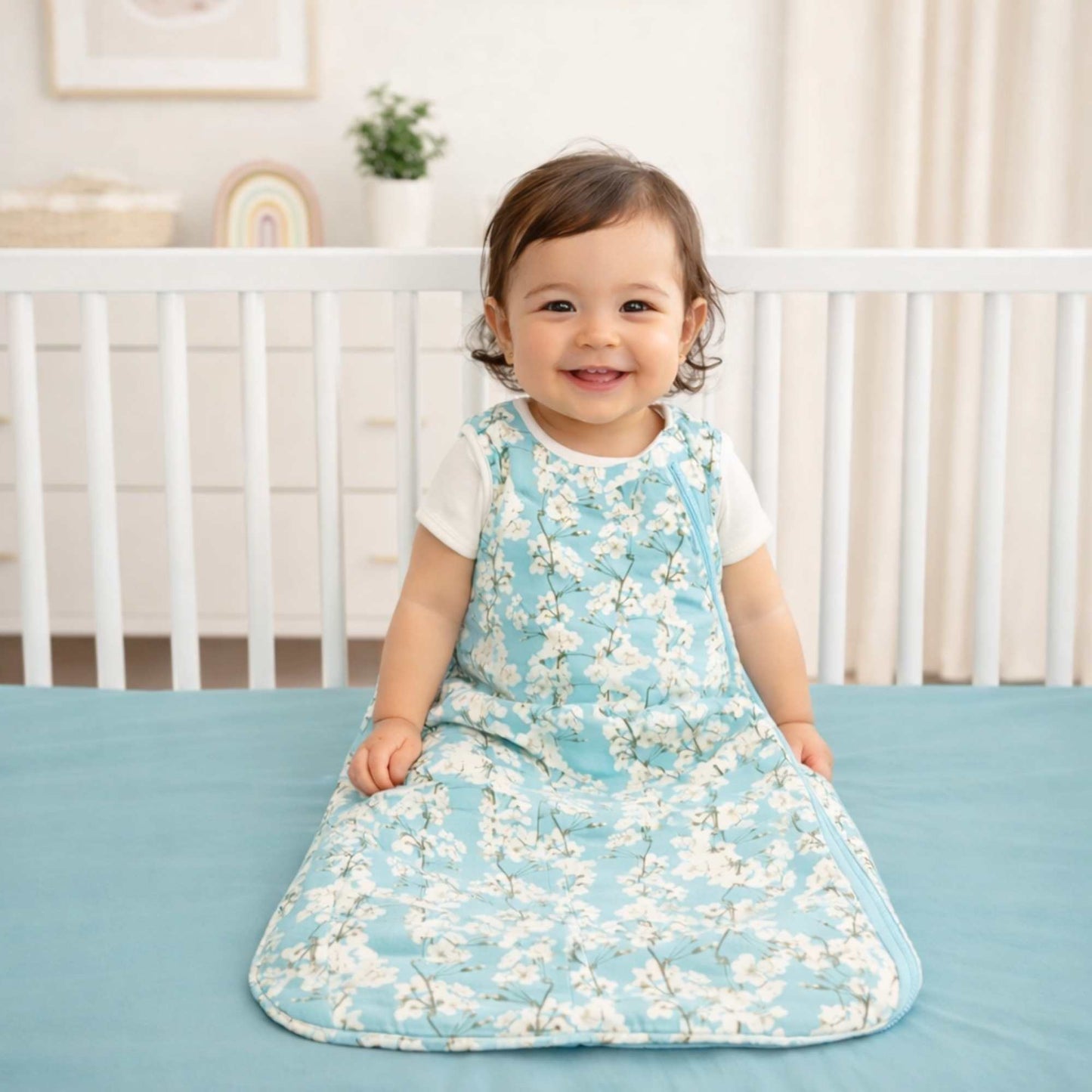 Child wearing a cherry blossom sleep sack by norani baby sitting on a crib in a nursery smiling. 