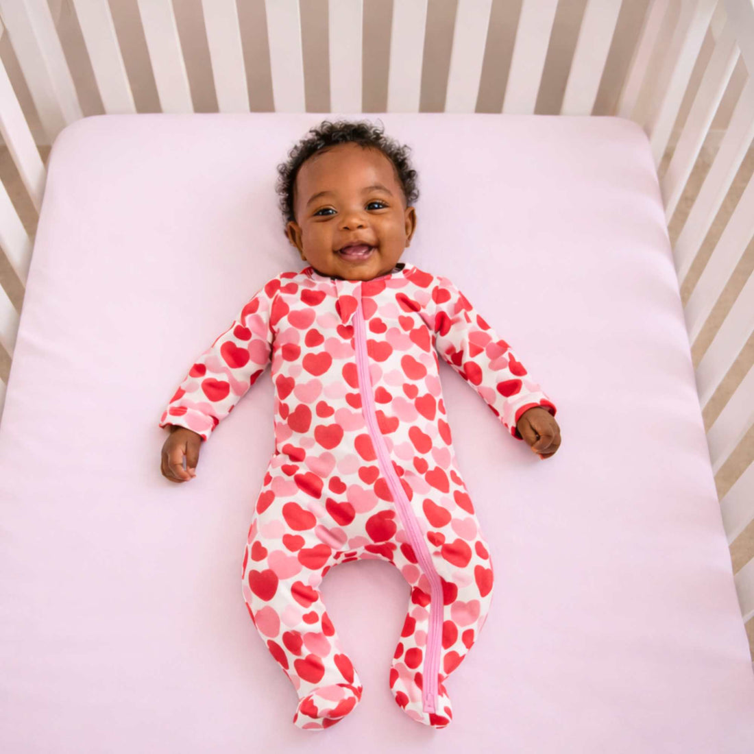 Baby wearing a pink onesie with red hearts by Norani Baby lying  in a crib.