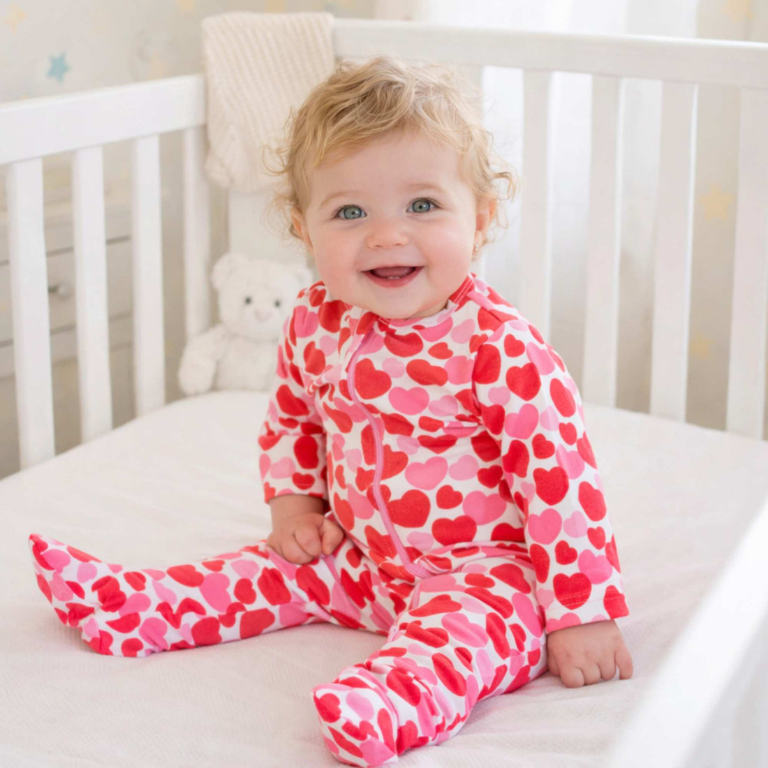 Child wearing a pink heart-patterned onesie sitting in a crib.