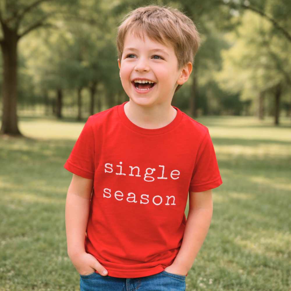 Kid outdoors wearing a bright red “Single Season” T-shirt, captured mid-movement with a playful smile, natural light, and a casual everyday setting.