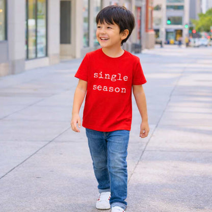 Kid walking down the street wearing a bright red “Single Season” T-shirt, smiling naturally in daylight with an urban outdoor backdrop.
