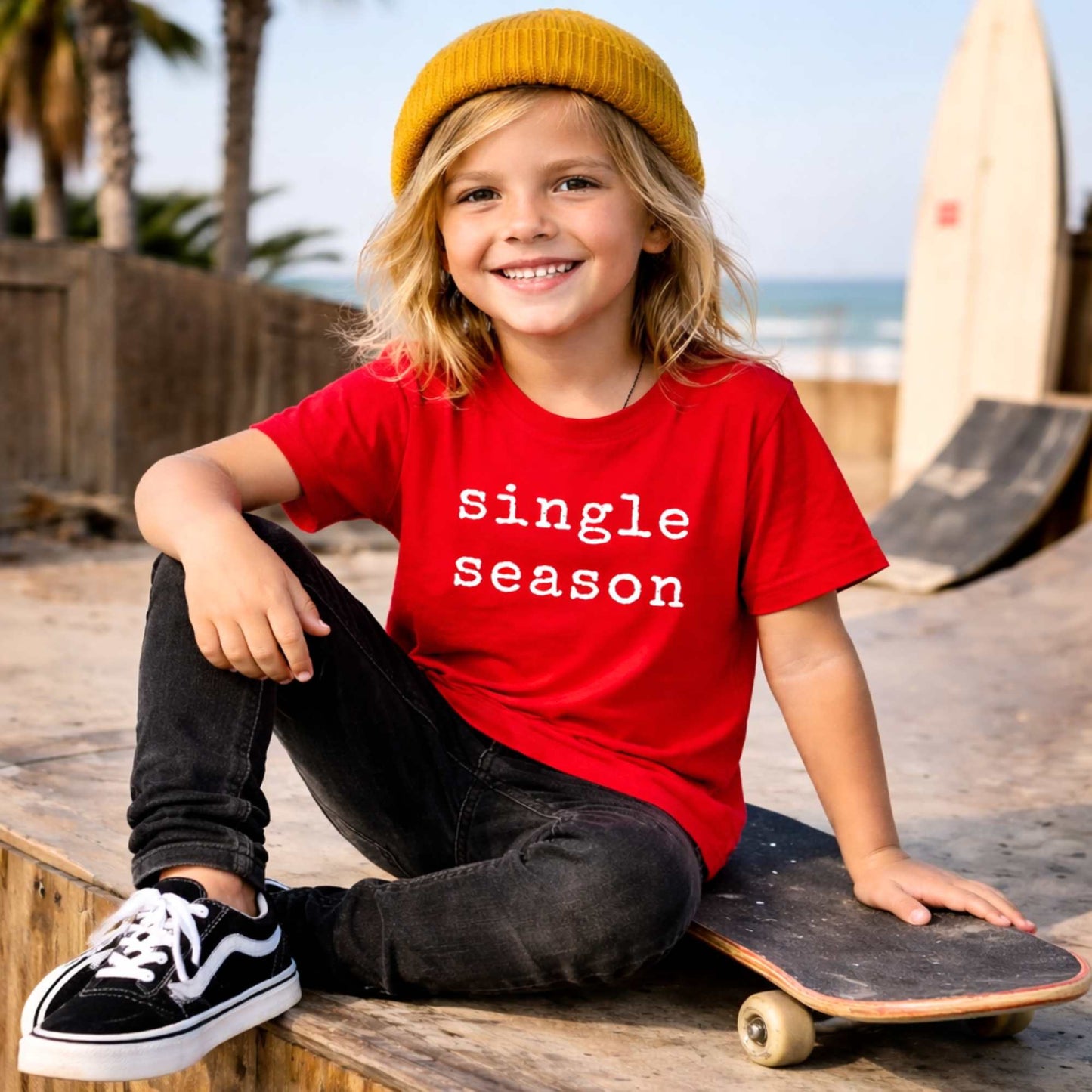 Child wearing a red shirt with 'single season' text, sitting on a skateboard at a skate park.