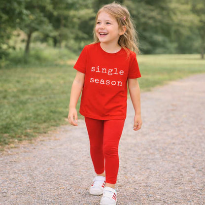 Girl walking through a park wearing a bright red “Single Season” T-shirt, smiling naturally in daylight with trees and open green space in the background.