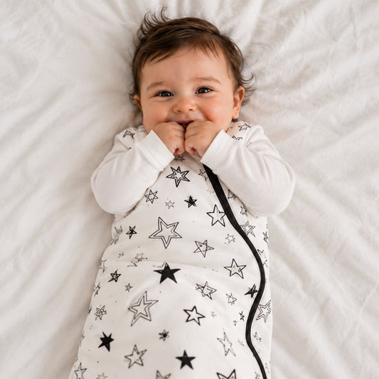 A baby lying on a white textured blanket, wearing a white long-sleeve swaddle wrap decorated with a black and gray star pattern and a black zipper.