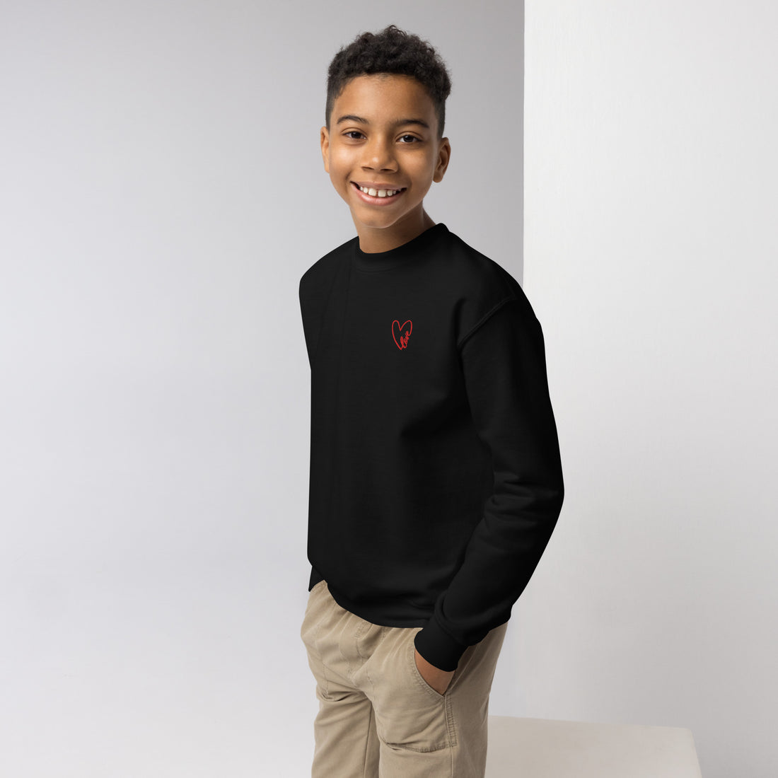 Boy smiling confidently with hands in his pockets, wearing a cozy crewneck sweatshirt featuring a small embroidered heart on the chest, styled for a casual, everyday look.