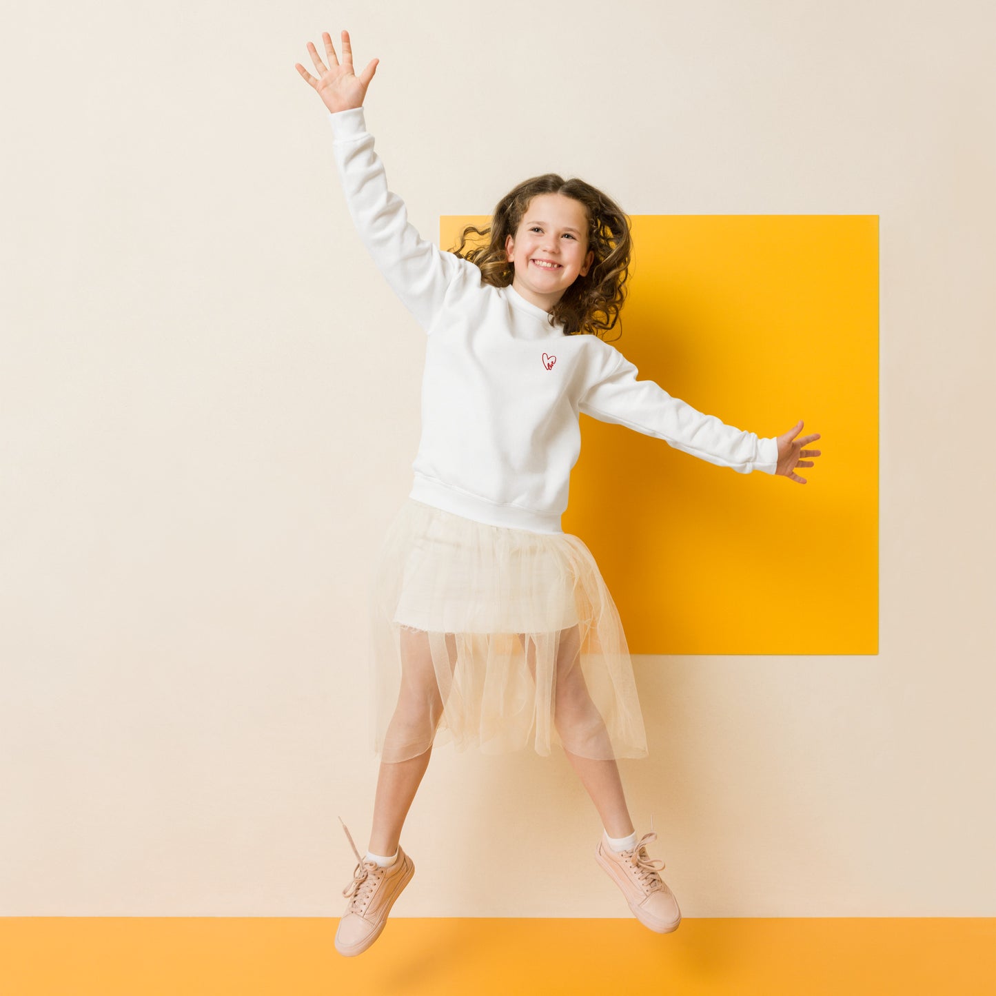 Girl mid-jump, smiling and full of energy, wearing a cozy crewneck sweatshirt with a small embroidered heart on the chest, captured in motion for a playful, everyday look.